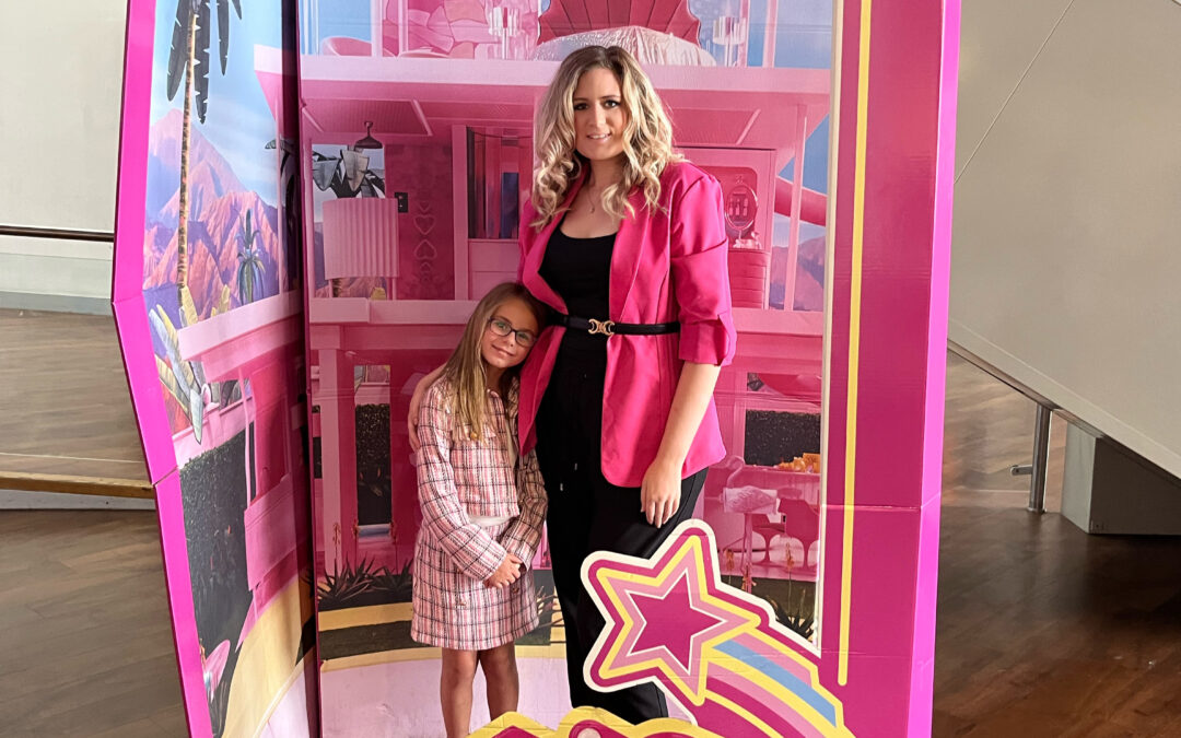 A woman in a bright pink blazer stands with her daughter inside a Barbie-themed life-size doll box display. Both are smiling, styled in pink outfits, symbolizing empowerment and identity.