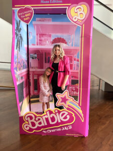 A woman in a bright pink blazer stands with her daughter inside a Barbie-themed life-size doll box display. Both are smiling, styled in pink outfits, symbolizing empowerment and identity.