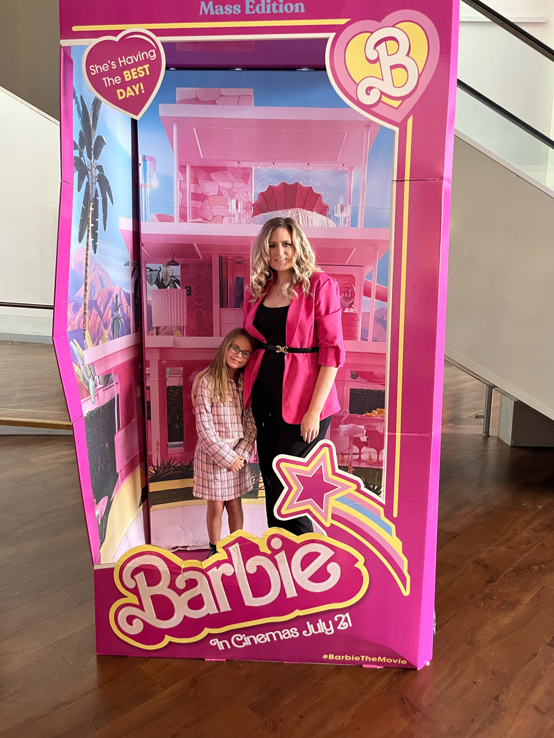 A woman in a bright pink blazer stands with her daughter inside a Barbie-themed life-size doll box display. Both are smiling, styled in pink outfits, symbolizing empowerment and identity.